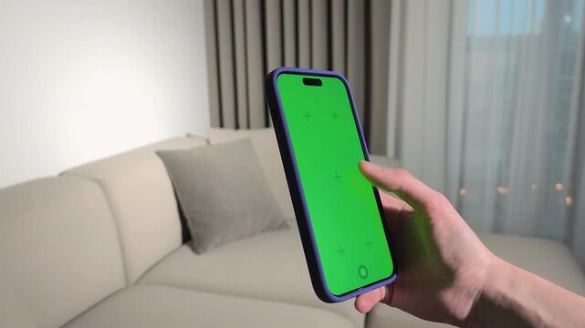 Hand holds smartphone with green screen in a living room. Finger taps the vertical mobile display in close up. App mockup for advertising and design..