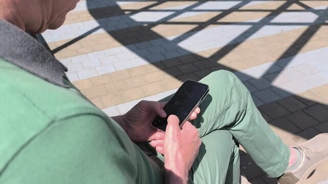 Man using smartphone outdoors in bright sunlight. Close up hands tap and scroll on mobile device over patterned pavement. Digital communication and browsing concept..