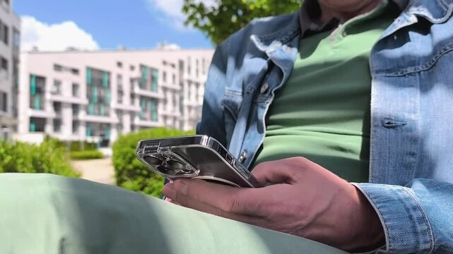 Man using smartphone outdoors in sunny courtyard. He taps and scrolls on a mobile screen beside modern apartments. Casual digital communication and browsing concept..