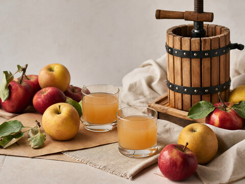 Several apples on a table next to two glasses of apple juice. A wooden apple press is also present, along with a cloth and some paper. The setting is a home kitchen.