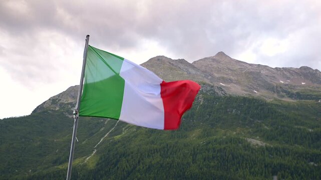 Italian flag waving in alpine mountain landscape with cloudy sky, summer travel destination in Italy, static low angle shot with copy space, tourism and patriotism concept