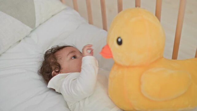Girl hispanic infant reaches with tiny hand to touch yellow plush duck beak while lying in a crib on soft bedding in a home nursery; curiosity.
