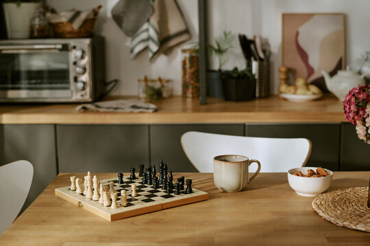 Wide shot of chessboard standing on kitchen table beside coffee mug and snack bowl, suggesting strategic thinking and leisure. Useful for lifestyle, home routine, planning, or hobby marketing
