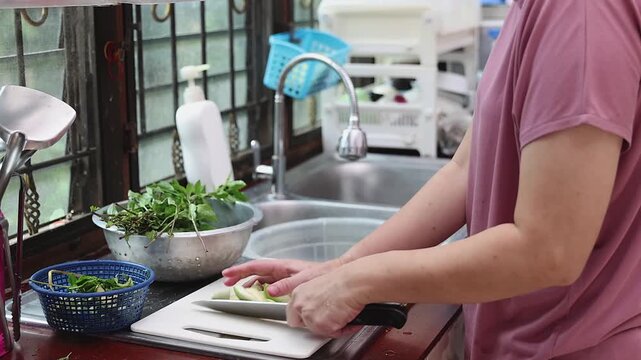 Side view of a housewife using a knife to slice fresh green Thai eggplants on a cutting board at the kitchen counter.
