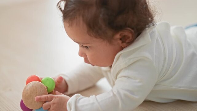 Girl chewing a wooden rattle with small hands on studio floor, mouth on teether while lying on tummy; curiosity play.