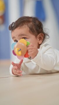 Hispanic baby girl on tummy reaching with hand for a colorful wooden rattle on floor, wide eyes and open mouth, crawling in a bright nursery; curiosity playfulness.