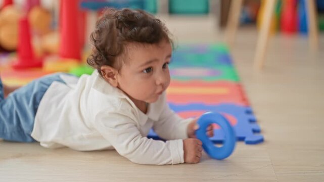 Girl chewing blue ring toy and reaching with one hand on colorful foam playroom mat near scattered toys and chair legs; curiosity.