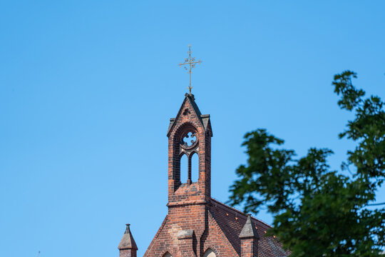 A brick church roof and small cross stand beneath a clear blue sky. Leafy deciduous trees frame the historic religious architecture.