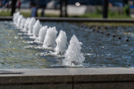A row of fountain jets bubbles across a shallow pool in bright sunlight. Soft background blur highlights the sparkling texture and motion of the water.