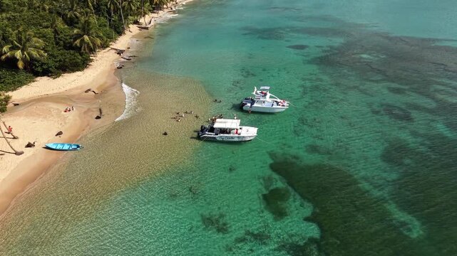 Aerial drone footage of Playa Coson coastline featuring turquoise ocean water, white sand beach, excursion boats and tropical palms in bright sunny weather.