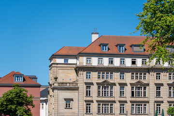 Fototapeta premium Historic stone facade with red tile roof stands under a clear blue sky. Green deciduous tree foliage frames the urban architecture.