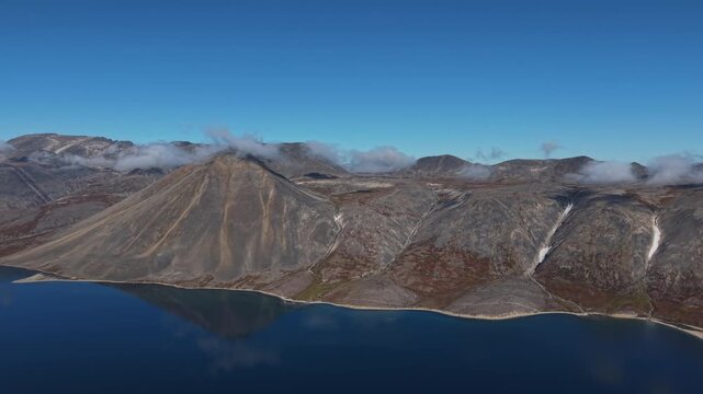 Aerial view orbits around a tall dramatic conical scree peak with autumn tundra slopes rising above the calm mirrored waters of pristine Abolesheva Bay on the remote arctic coast of Chukotka at midday