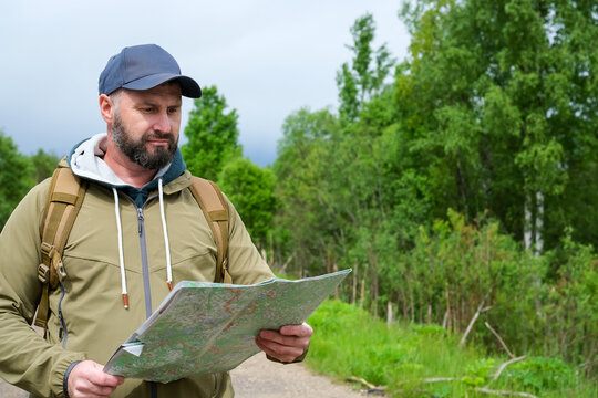Bearded man with backpack reading paper map on rural forest road under cloudy sky during outdoor trip