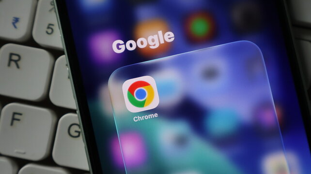 Closeup of a smartphone screen displaying the google chrome app icon inside a folder on a blurred background, next to a computer keyboard