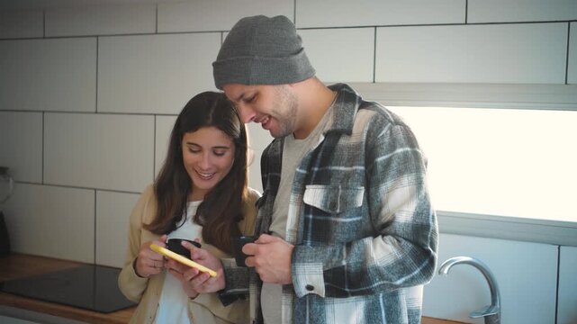 Video of a young Caucasian couple on a winter day in their home kitchen enjoying a cup of coffee while watching videos on their mobile phone. Concept of a winter day at home, young marriage, and famil