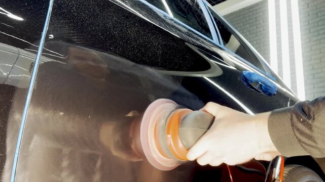 A car service worker polishes the car body with a polishing machine. Detailing and polishing of the car. Car detailing - hands with an orbital polishing machine in an auto repair shop. 