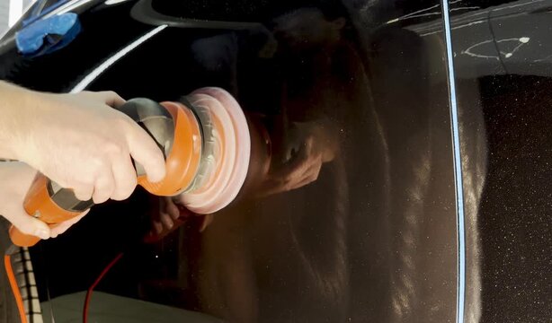 A car service worker polishes the car body with a polishing machine. Detailing and polishing of the car. Car detailing - hands with an orbital polishing machine in an auto repair shop. 