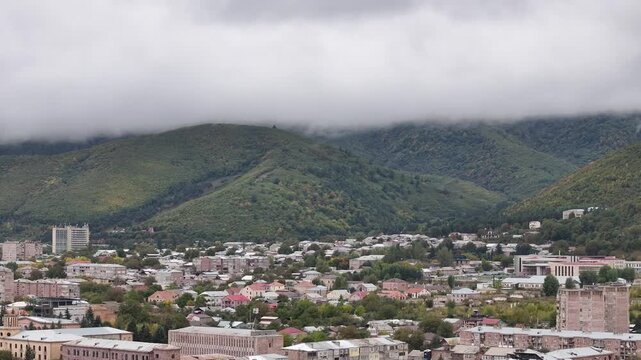 Wide aerial view of a scenic mountain valley city with residential buildings and green trees under a dramatic, cloudy sky with thick fog rolling over the forested hills.

