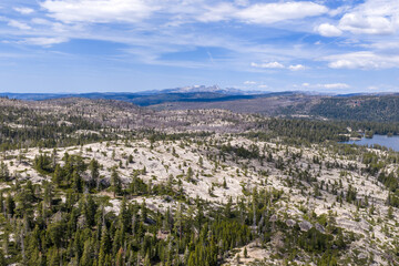 Obraz premium Wide landscape showcasing rugged granite rock formations, sparse pine trees, and a distant shimmering mountain lake under a bright blue sky with scattered clouds