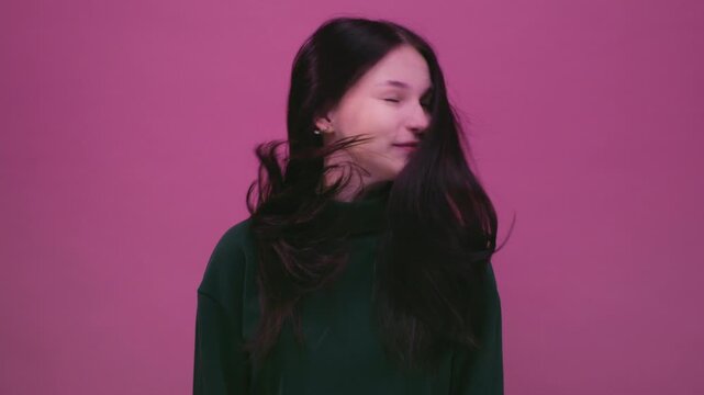 Beautiful cheerful brunette young girl wearing green pink sweater posing isolated on pink wall. Dancing shaking head with flowing hair. Teenage woman shakes her straight black long hair, slow motion.