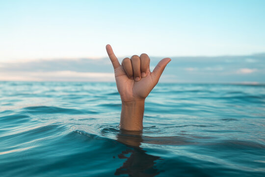 Human hand performing shaka sign gesture emerging from blue ocean water