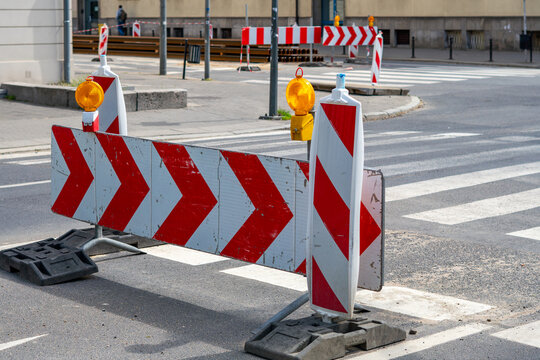 Road barriers with white and red stripes and warning signs with chevrons and yellow lamps, placed on asphalt near a pedestrian crossing during public construction works in the city, photographed in da