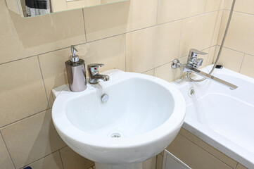 Fototapeta premium white pedestal sink with a chrome faucet and soap dispenser sits next to a bathtub with a flexible shower hose; beige tiles cover the walls