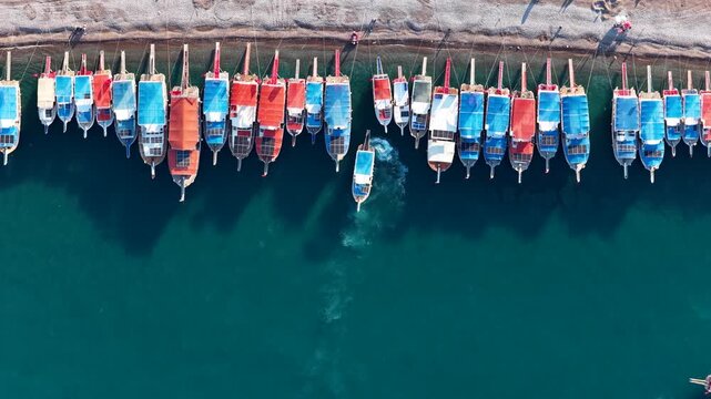 Top-down aerial drone view of traditional colorful boats moored along the turquoise coastline, creating a perfect geometric pattern on a sunny summer day.