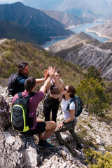 Group of friends hiking in the mountains celebrating with a high five on a rocky trail overlooking a turquoise lake and dramatic ridgelines © Nikola Spasenoski