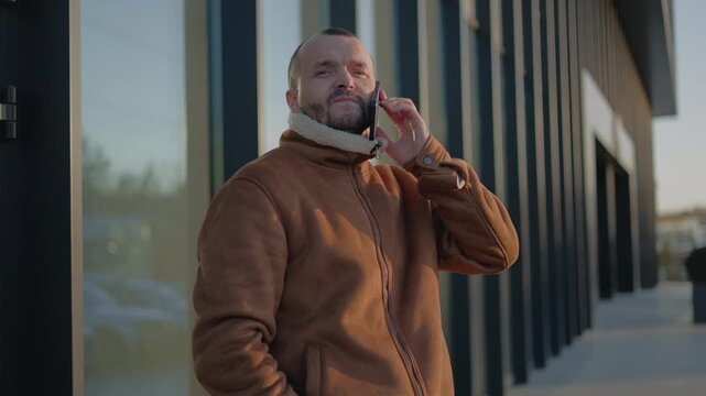 His facial expression shows positive emotions from the ongoing call.The general mood of the man appears satisfied and optimistic.He wears a warm jacket in a muted brown shade.
