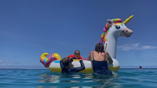 Two women and two boys playing on inflatable unicorn float in turquoise water under clear blue sky, friends laughing and splashing, lounging on giant horned float, sunny relaxed, Bali island
