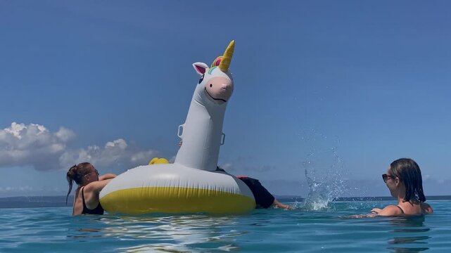 Two women and two boys playing on inflatable unicorn float in turquoise water under clear blue sky, friends laughing and splashing, lounging on giant horned float, sunny relaxed, Bali island