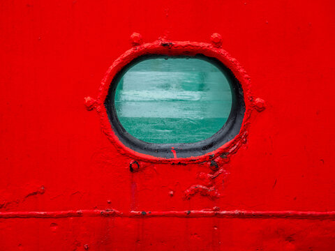 Oval ship porthole window on bright red metal hull background.  Close-up of a classic nautical porthole on a vessel side painted in vibrant red. Through the glass, green deck or sea surface is visible
