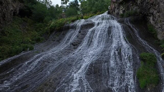 A stunning wide shot of the majestic cascading mountain waterfall flowing down a dark rocky cliff surrounded by lush green vegetation on a beautiful summer day in Armenia.

