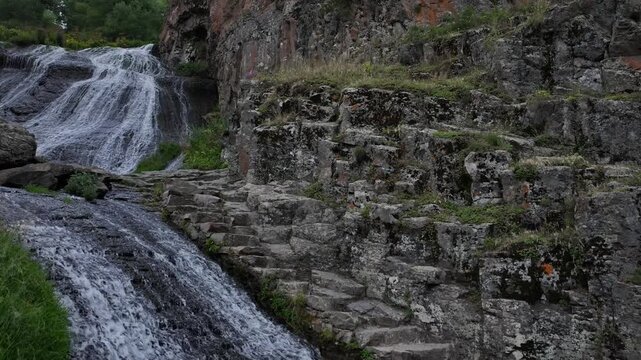 A stunning wide shot of the majestic cascading mountain waterfall flowing down a dark rocky cliff surrounded by lush green vegetation on a beautiful summer day in Armenia.

