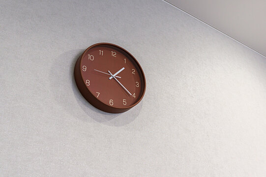 A matte brown round wall clock with white numerals and hands is mounted on a light grey textured wall