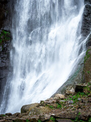 Obraz premium Powerful mountain waterfall spraying over wet dark rocks. White water spray and foam contrast with wet dark rocks and green moss, creating a dynamic nature background.