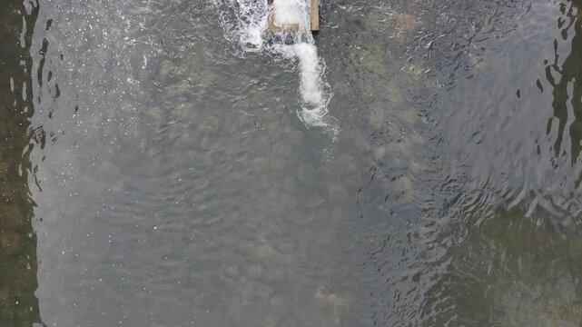 An aerial top-down shot of water cascading over a small concrete dam structure on a river with rocks visible through the clear water.

