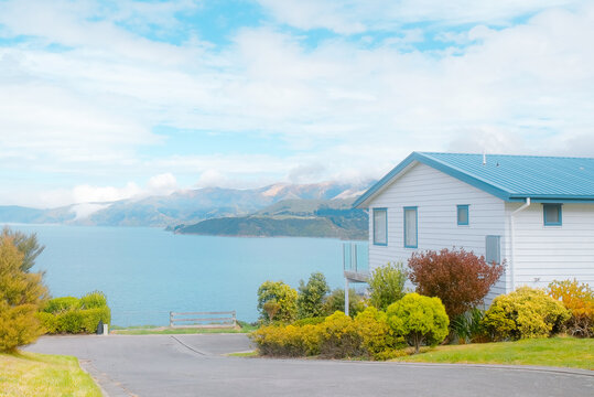 South Island, New Zealand: charming white holiday home with blue roof, vacation house with calm turquoise sea bay and distant volcanic mountain view.