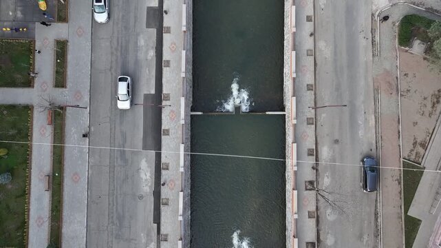 An aerial top-down shot of water cascading over a small concrete dam structure on a river with rocks visible through the clear water.

