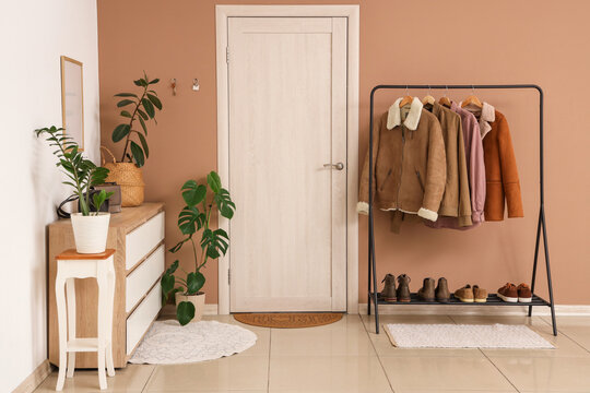 Cozy modern entryway interior with coat rack, autumn jackets, shoes, houseplants and wooden door in warm beige hallway at home