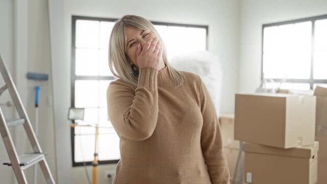 Woman with hand covering mouth laughing among cardboard boxes inside a building; joyful moving day now.