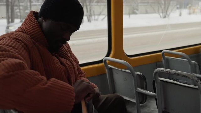 Black man adjusting jacket and bag on snowy bus, fastening zipper, checking watch, searching pocket for ticket, peering out window at frosty roadway, wearing orange puffer and beanie, breath fog