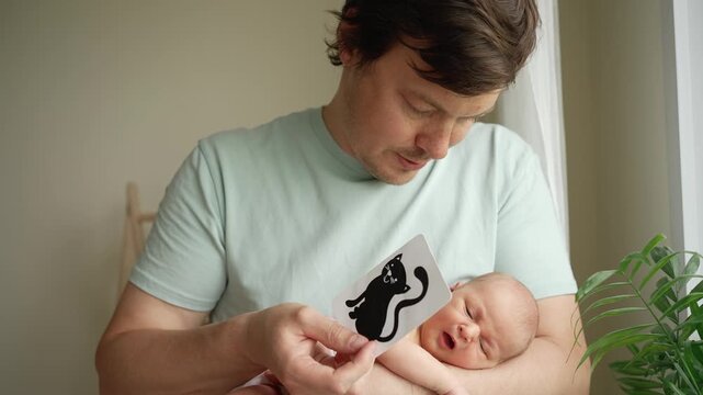 Young caring father holding his newborn baby in his arms while showing him high contrast black and white flashcards with animal pictures, aiding in early visual stimulation