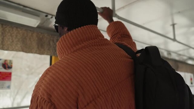 Man back facing bus window wearing chunky orange knit, black beanie, backpack straps visible subtle city posters, muted light, solitude and travel fatigue student returning home after long day.