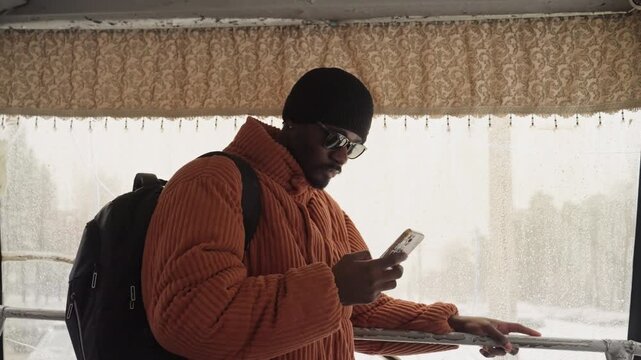Black student in orange puffer jacket checking smartphone while waiting inside bus shelter, snow falling against window, backpack straps visible, reflective glass, moody warm tones, introspective