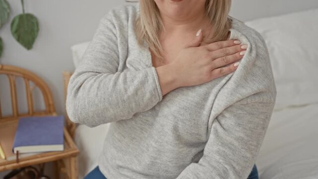 Woman, blonde, clutching chest with hand on chest gesture suggesting heartattack while seated on bed beside a book and rattan chair in studio; concern health awareness.