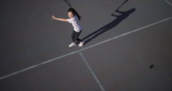 A young girl with focused intensity plays tennis, her dynamic pose captured as she reaches for the ball on an outdoor court under clear skies.