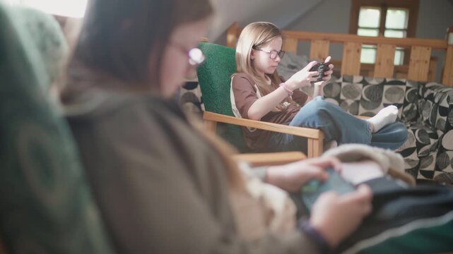 Two Sisters Sitting in Comfortable Chair in Kids Room Cheerfully Smiling and Laughing While Playing Online Game Together Using Smartphones Connected to Internet Happy Children Game Addiction Concept