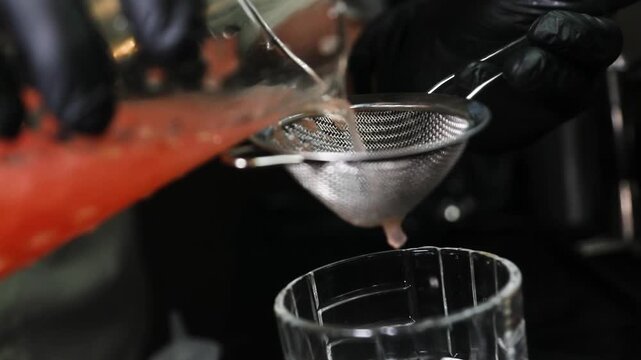Barman colando l&iacute;quido con colador met&aacute;lico sobre fondo negro. Barman profesional filtrando un c&oacute;ctel con un colador met&aacute;lico. C&oacute;ctel artesanal, t&eacute;cnica de bar precisa, servicio de primera.